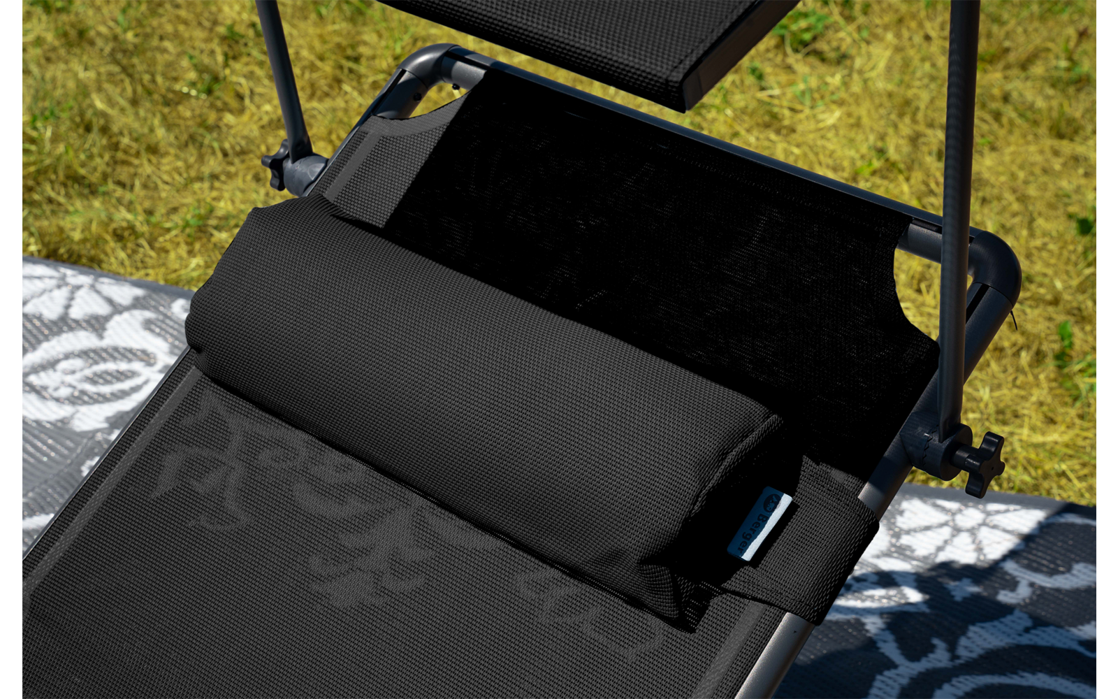 Schwarzer Liegestuhl mit gepolsterter schwarzer Nackenrolle, Befestigungsriemen und Metallstangen mit Gelenken, auf einer grauen Matte mit Blumenmuster auf Gras