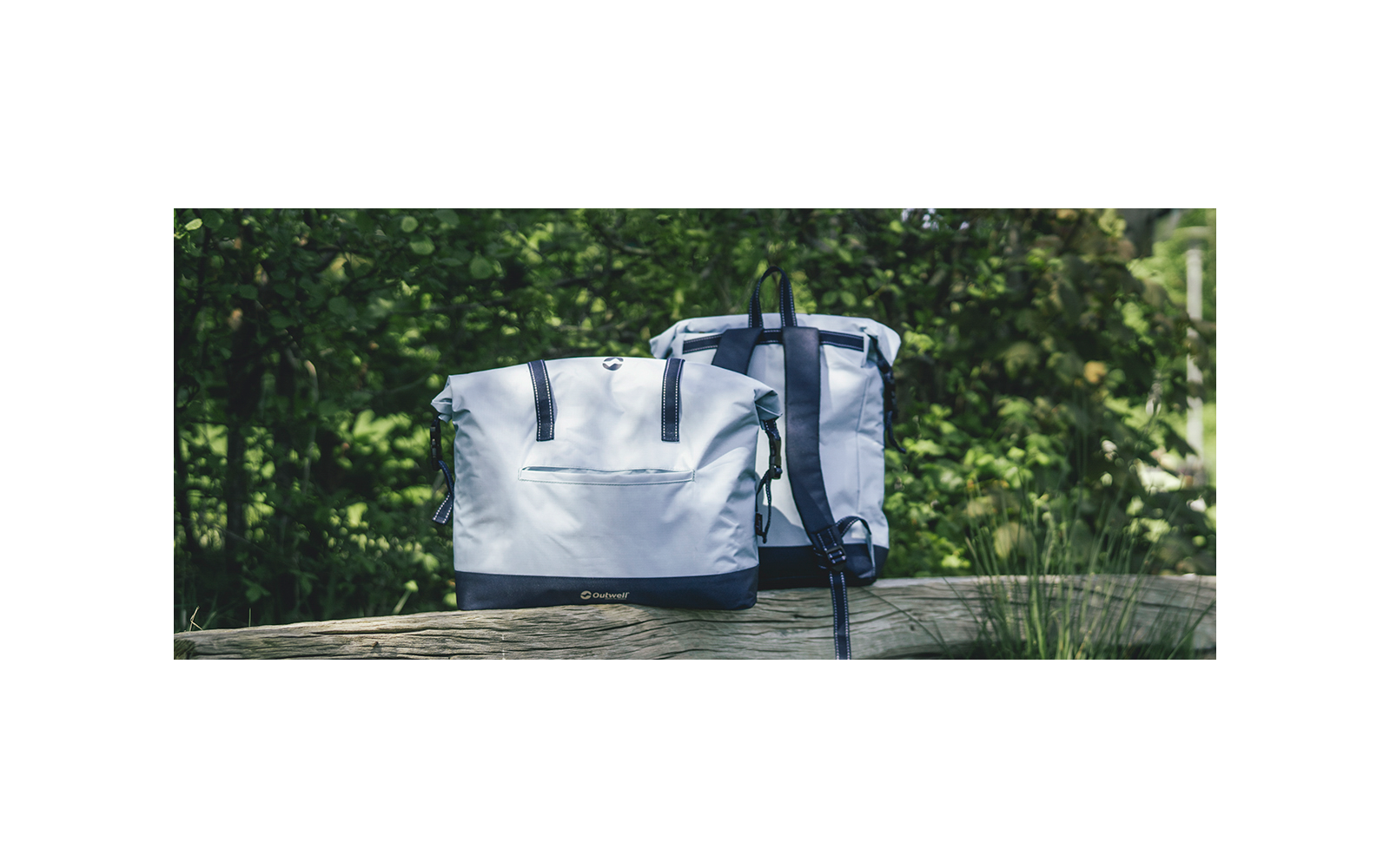 Zwei hellgraue Rucksäcke auf einer Holzbank, links mit Fronttasche, rechts mit sichtbaren Schultergurten; grünes Laub im Hintergrund