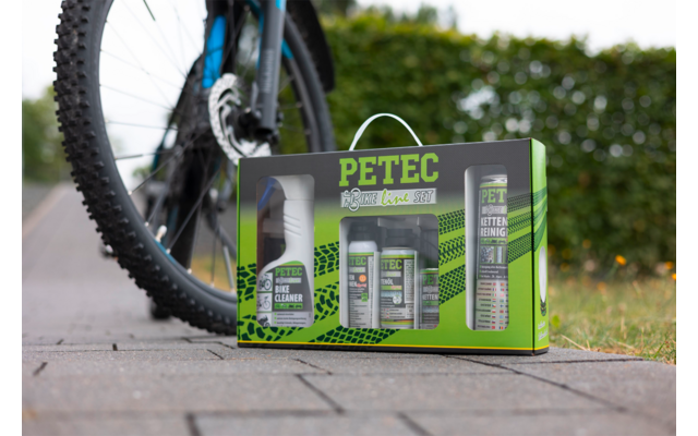 Groene PETEC-doos met meerdere spuitbussen en flessen op bestrating naast het achterwiel van een fiets, haag op de achtergrond