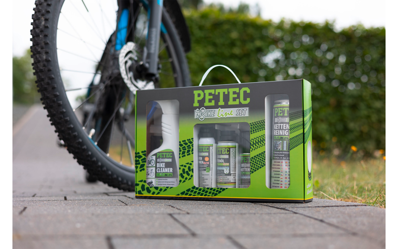 Groene PETEC-doos met meerdere spuitbussen en flessen op bestrating naast het achterwiel van een fiets, haag op de achtergrond