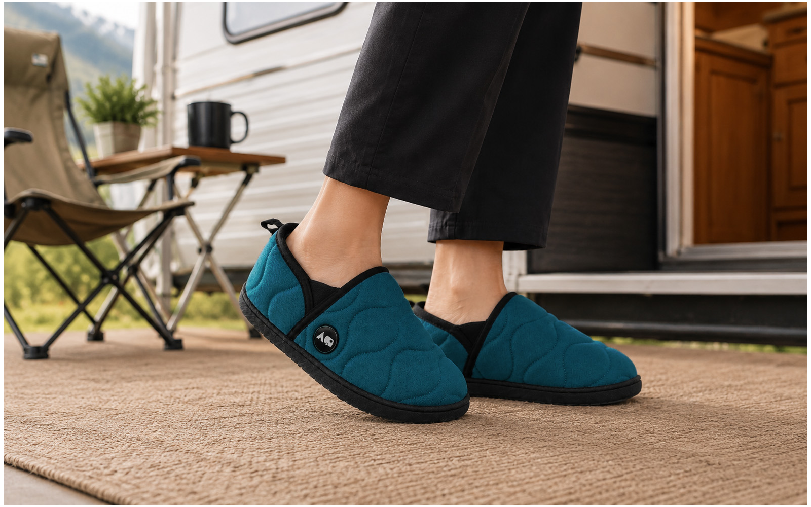 Pieds en chaussons turquoise à bordure noire sur un paillasson tissé devant la porte ouverte d'une caravane ; chaise de camping pliante, petite table avec tasse noire et pot de plante, montagnes floues en arrière-plan