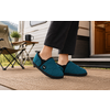 Pieds en chaussons turquoise à bordure noire sur un paillasson tissé devant la porte ouverte d'une caravane ; chaise de camping pliante, petite table avec tasse noire et pot de plante, montagnes floues en arrière-plan