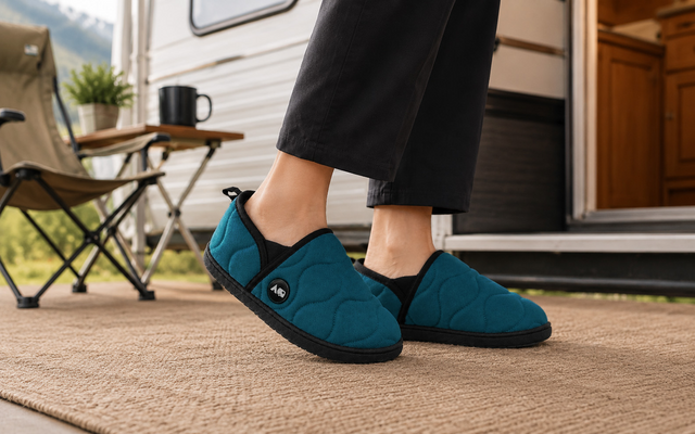 Pieds en chaussons turquoise à bordure noire sur un paillasson tissé devant la porte ouverte d'une caravane ; chaise de camping pliante, petite table avec tasse noire et pot de plante, montagnes floues en arrière-plan