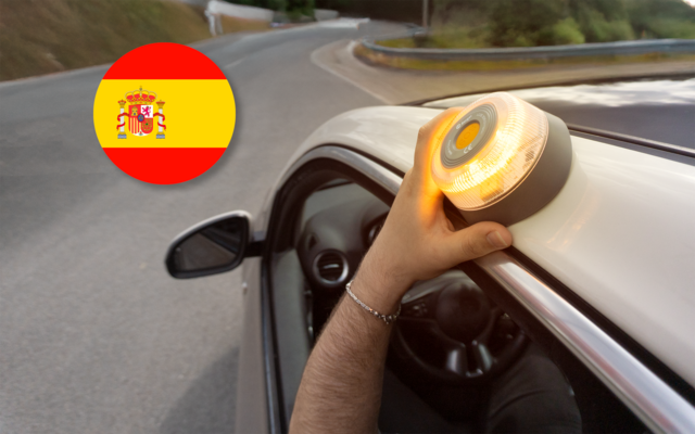 Hand mit Armband hält eine gelbe runde Leuchte auf dem Autodach; Blick aus dem Innenraum über Lenkrad und Außenspiegel auf eine kurvige Straße mit Leitplanke; oben links ein kreisförmiges Symbol mit spanischer Flagge