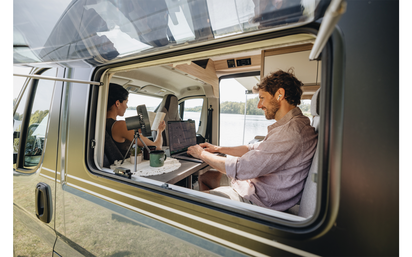 Blick durch geöffnetes Wohnmobilfenster auf zwei Personen am Tisch: eine Person tippt auf einem Laptop, die andere hält eine Kamera; Laptop, grüner Becher, See und Wiese im Hintergrund