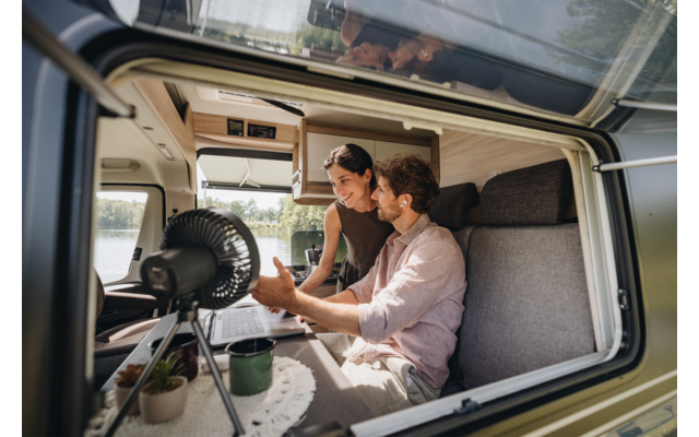 Zwei Personen in einem Wohnmobil: ein Mann sitzt auf der Bank, eine Frau lehnt sich über ihn; auf dem Tisch stehen ein Laptop, eine Tasse, ein kleiner Ventilator und eine Topfpflanze; durch das Fenster sind See und Bäume sichtbar