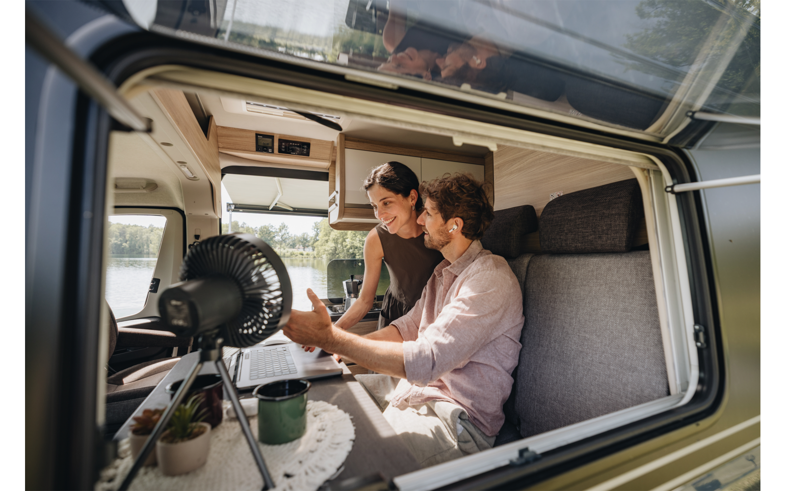 Zwei Personen in einem Wohnmobil: ein Mann sitzt auf der Bank, eine Frau lehnt sich über ihn; auf dem Tisch stehen ein Laptop, eine Tasse, ein kleiner Ventilator und eine Topfpflanze; durch das Fenster sind See und Bäume sichtbar