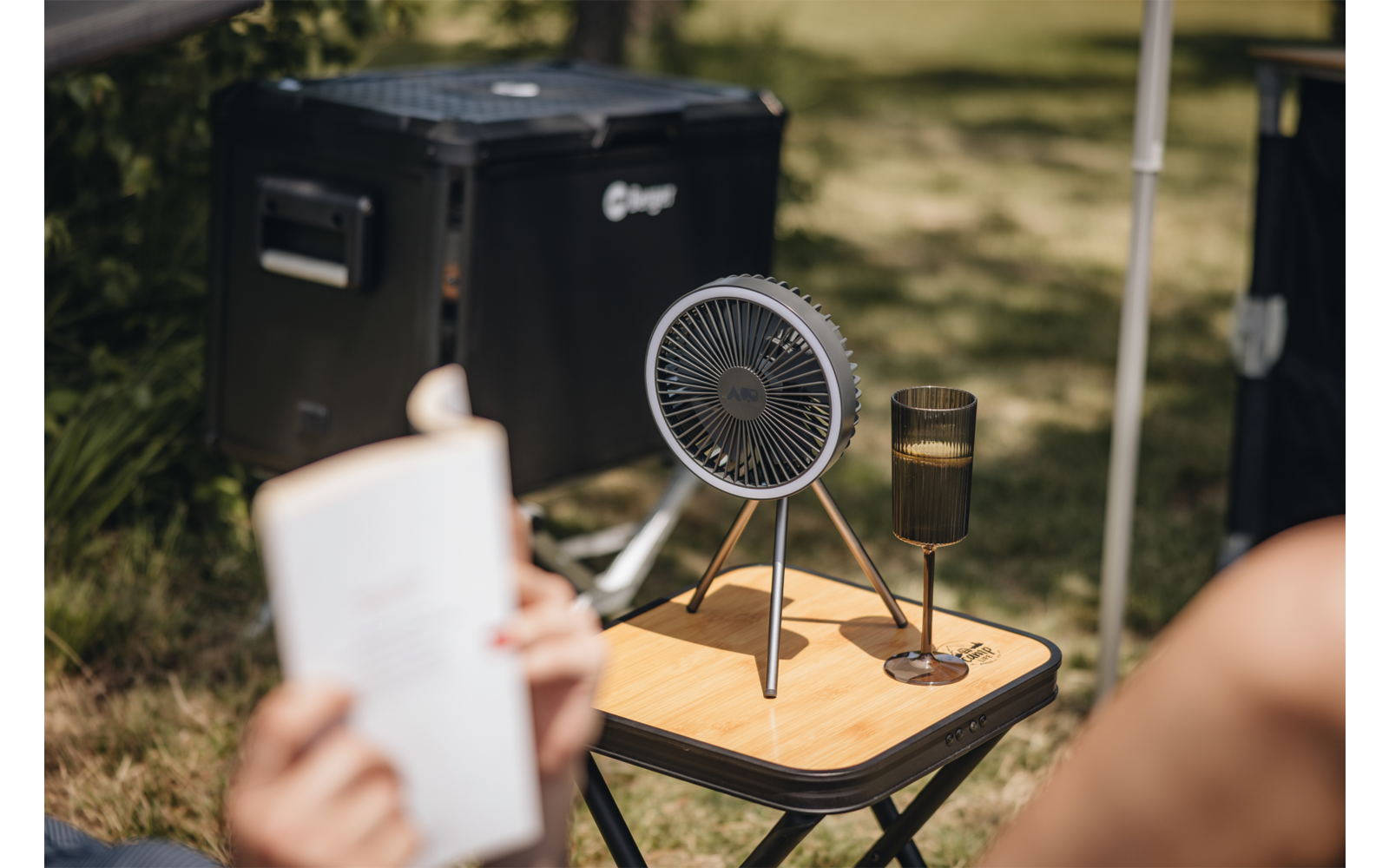 Kleiner tragbarer Ventilator auf einem kleinen Klapptisch aus Holz, daneben ein Stielglas mit Getränk; im Vordergrund unscharfe Hände, die ein aufgeschlagenes Buch halten; im Hintergrund eine schwarze Campingkiste und Rasen