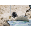 Kleiner tragbarer Ventilator auf Dreibein steht auf Sand neben einer Strandmatte mit Zickzackmuster und einem gehäkelten Kissen; im Hintergrund ein Liegestuhlbein und ein Sonnenschirmstiel