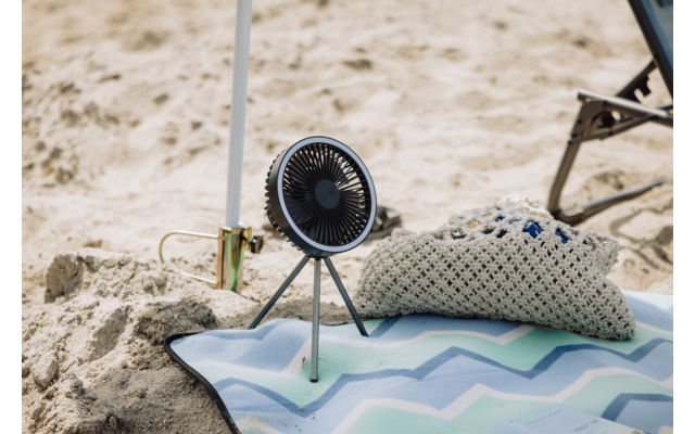 Kleiner tragbarer Ventilator auf Dreibein steht auf Sand neben einer Strandmatte mit Zickzackmuster und einem gehäkelten Kissen; im Hintergrund ein Liegestuhlbein und ein Sonnenschirmstiel