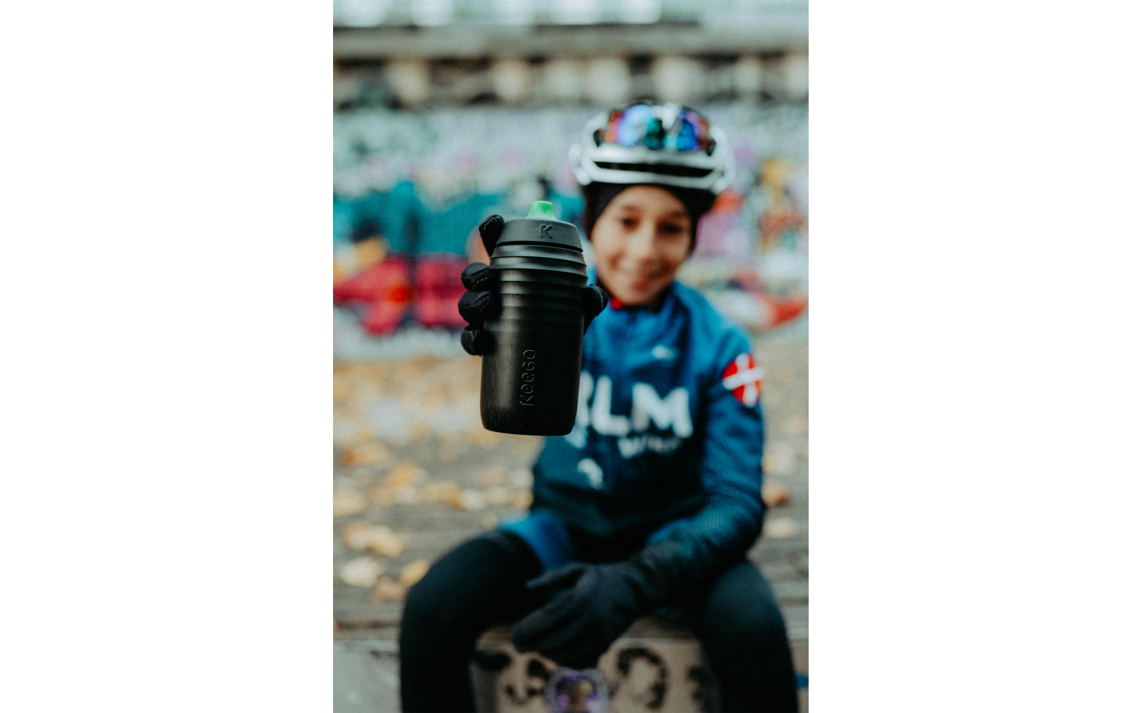 Een kind met fietshelm en handschoenen houdt een zwarte drinkfles naar de camera; onscherpe achtergrond, gevallen bladeren op de grond