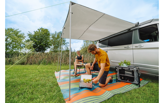 Twee personen onder een luifel naast een geparkeerde camper, één persoon knielt bij een krat met etenswaren, de ander zit op een kruk op een kleurrijk kleed; draagbare barbecue, gras en bomen zichtbaar