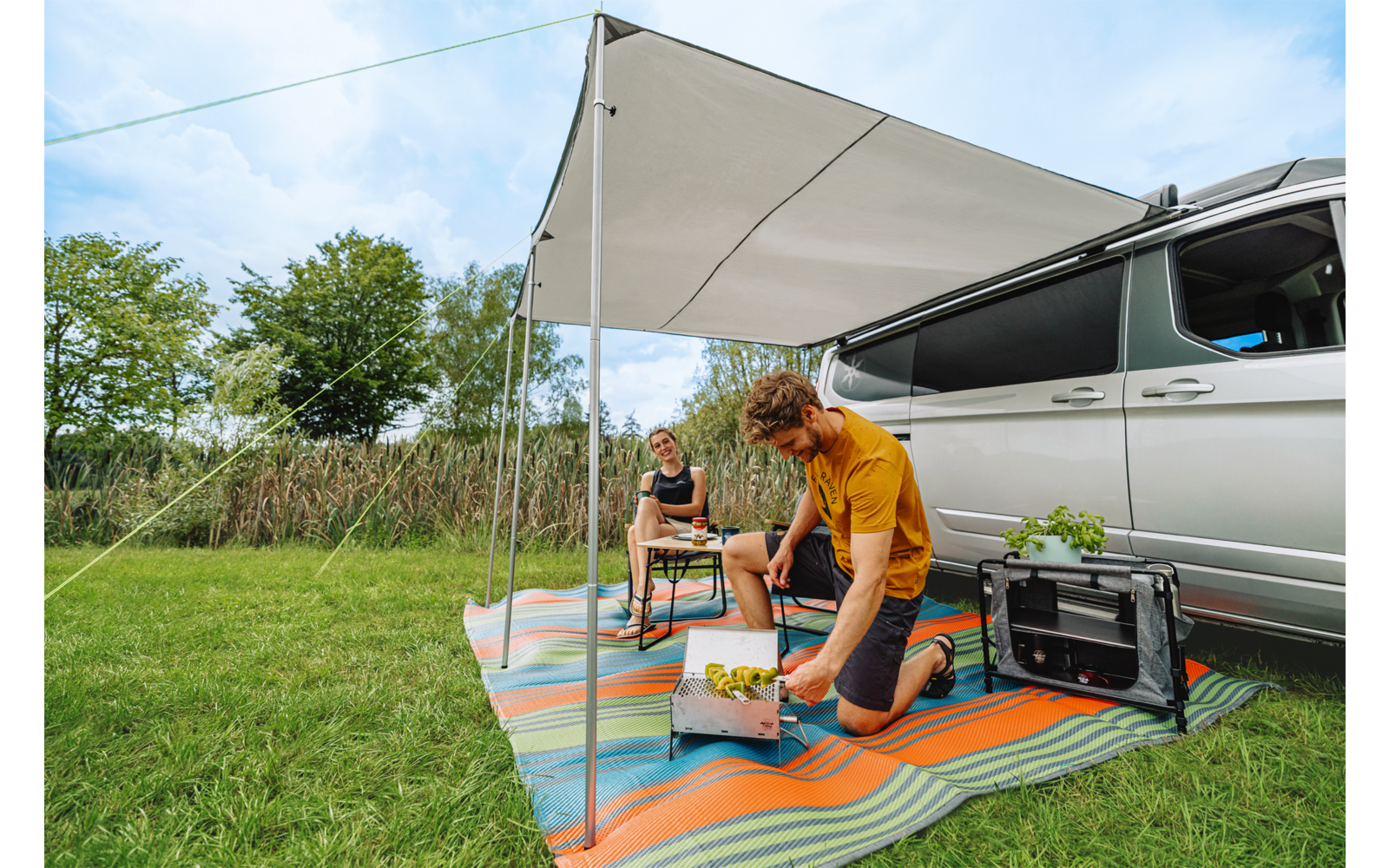 Twee personen onder een luifel naast een geparkeerde camper, één persoon knielt bij een krat met etenswaren, de ander zit op een kruk op een kleurrijk kleed; draagbare barbecue, gras en bomen zichtbaar
