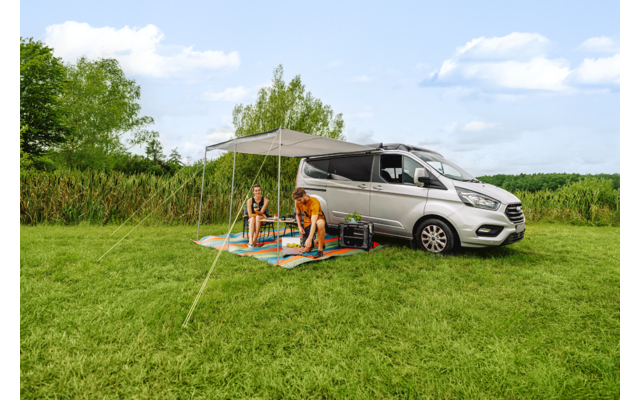 Grijs voertuig met uitgeschoven luifel op een grasveld; twee personen zitten op kleurrijke matten onder de luifel; riet, bomen en een blauwe lucht met wolken op de achtergrond
