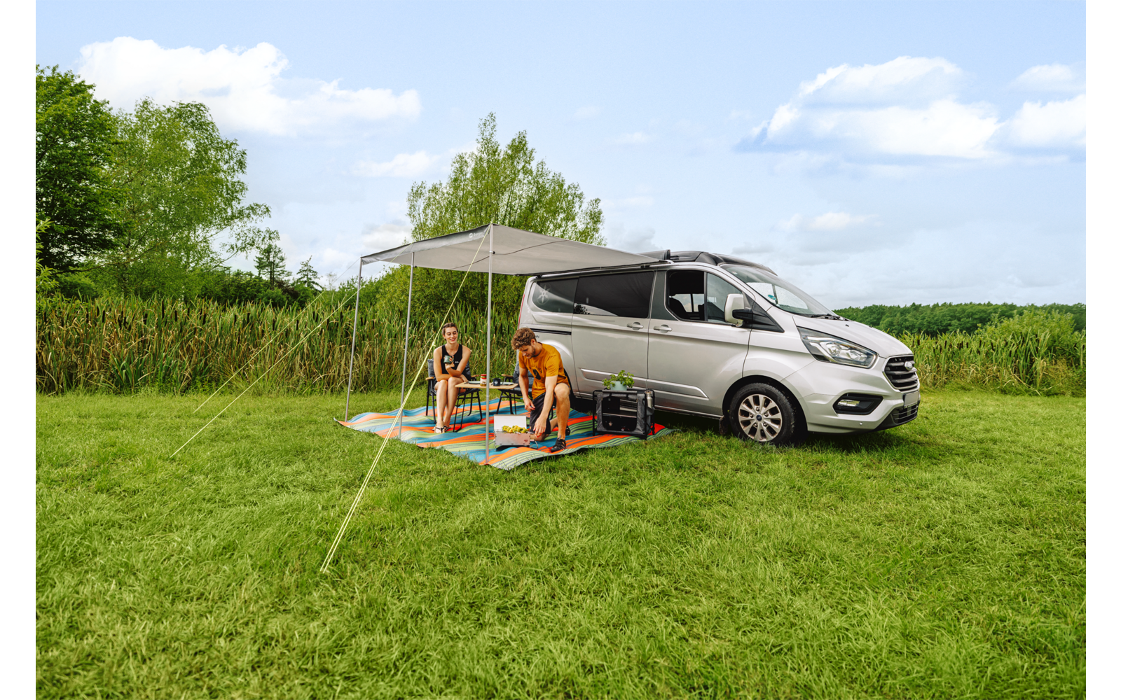 Grijs voertuig met uitgeschoven luifel op een grasveld; twee personen zitten op kleurrijke matten onder de luifel; riet, bomen en een blauwe lucht met wolken op de achtergrond
