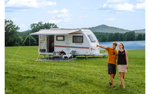 Caravan op een groene wei bij een meer; twee personen lopen naar rechts, één wijst; luifel uitgeklapt, inklapbare tafel en stoelen voor de caravan; beboste heuvels op de achtergrond