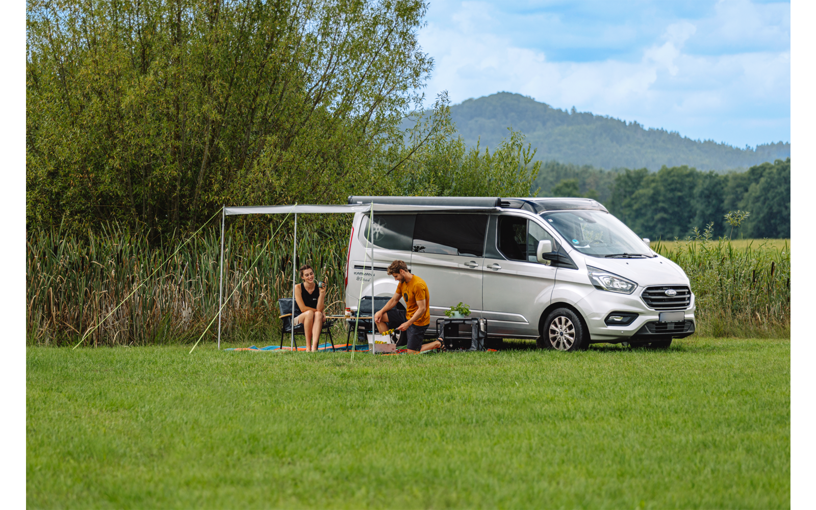 Twee personen zitten naast een geparkeerde witte camper met uitgeklapte luifel op een groen veld; riet, bomen en heuvels op de achtergrond