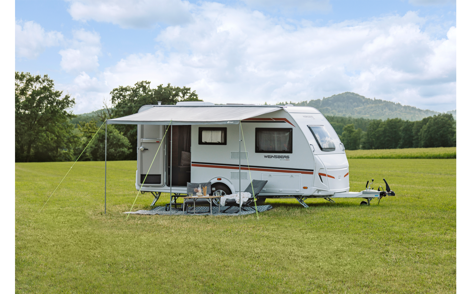 Witte caravan op een grasveld met een uitgeklapt zonnescherm, open deur, inklapbare tafel en stoelen; bomen en heuvels op de achtergrond onder een lichtbewolkte hemel