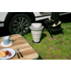 Gedeeltelijke weergave van een witte auto met wiel; persoon in zwarte broek stapt op een zwarte opstap; inklapbaar wit draagbaar toilet op gras; houten tafel met blauwe mok, bord met croissant en druiven en bestek