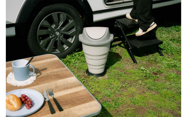 Gedeeltelijke weergave van een witte auto met wiel; persoon in zwarte broek stapt op een zwarte opstap; inklapbaar wit draagbaar toilet op gras; houten tafel met blauwe mok, bord met croissant en druiven en bestek