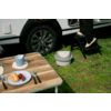 Tafel met blauwe mok op onderzetter, bord met croissant en rode druiven, bestek; op gras een grijs draagbaar toilet, een wiel van een wit voertuig en een persoon in zwarte broek met witte schoenen op een zwart opstapje