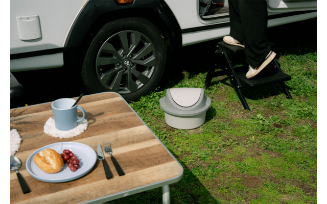 Tafel met blauwe mok op onderzetter, bord met croissant en rode druiven, bestek; op gras een grijs draagbaar toilet, een wiel van een wit voertuig en een persoon in zwarte broek met witte schoenen op een zwart opstapje