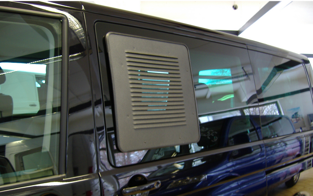 Véhicule utilitaire foncé, porte latérale coulissante avec grille d'aération rectangulaire, reflets sur la vitre, rangées de sièges visibles à travers la vitre