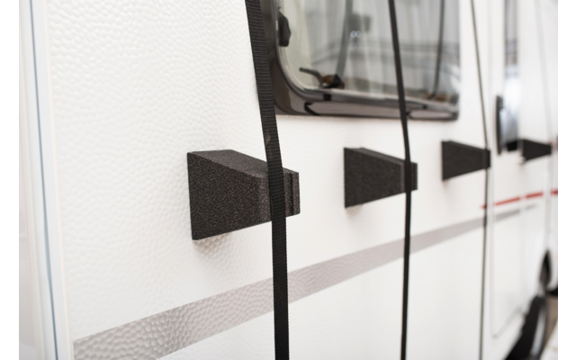 Weiße Seitenwand eines Fahrzeugs mit Fenster, drei schwarze keilförmige Blöcke mit hängenden schwarzen Gurten und einer grauen sowie dünnen roten Streifen
