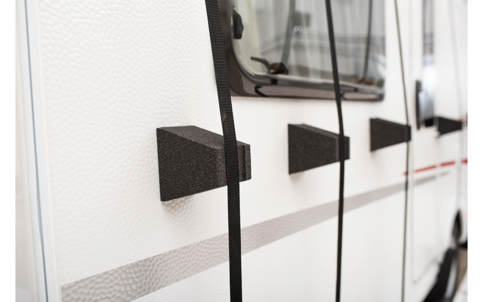 Weiße Seitenwand eines Fahrzeugs mit Fenster, drei schwarze keilförmige Blöcke mit hängenden schwarzen Gurten und einer grauen sowie dünnen roten Streifen