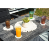 Mesa de madera oscura con cuatro vasos acanalados con líquido de color, cada uno sobre un posavasos redondo blanco; en el centro una maceta de cerámica blanca con planta verde sobre un tapete redondo blanco de ganchillo; al fondo hierba, parte de una silla y el borde de un vehículo blanco