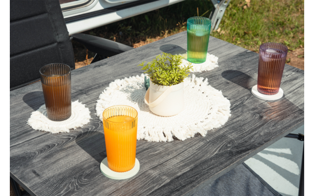 Mesa de madera oscura con cuatro vasos acanalados con líquido de color, cada uno sobre un posavasos redondo blanco; en el centro una maceta de cerámica blanca con planta verde sobre un tapete redondo blanco de ganchillo; al fondo hierba, parte de una silla y el borde de un vehículo blanco