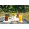 Mesa de madera gris al aire libre con cuatro vasos acanalados (marrón, verde, morado, naranja), una maceta blanca con planta verde sobre un paño blanco, césped y árboles al fondo