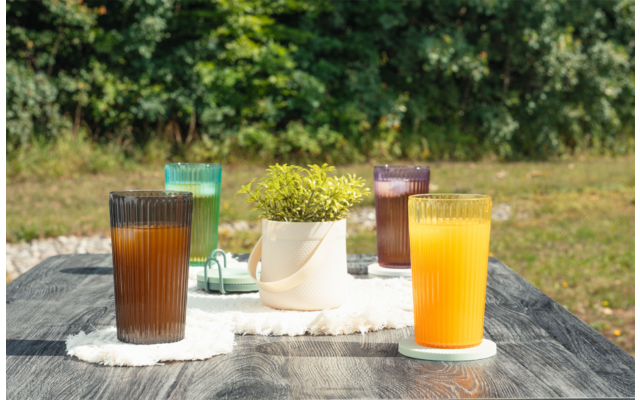 Mesa de madera gris al aire libre con cuatro vasos acanalados (marrón, verde, morado, naranja), una maceta blanca con planta verde sobre un paño blanco, césped y árboles al fondo