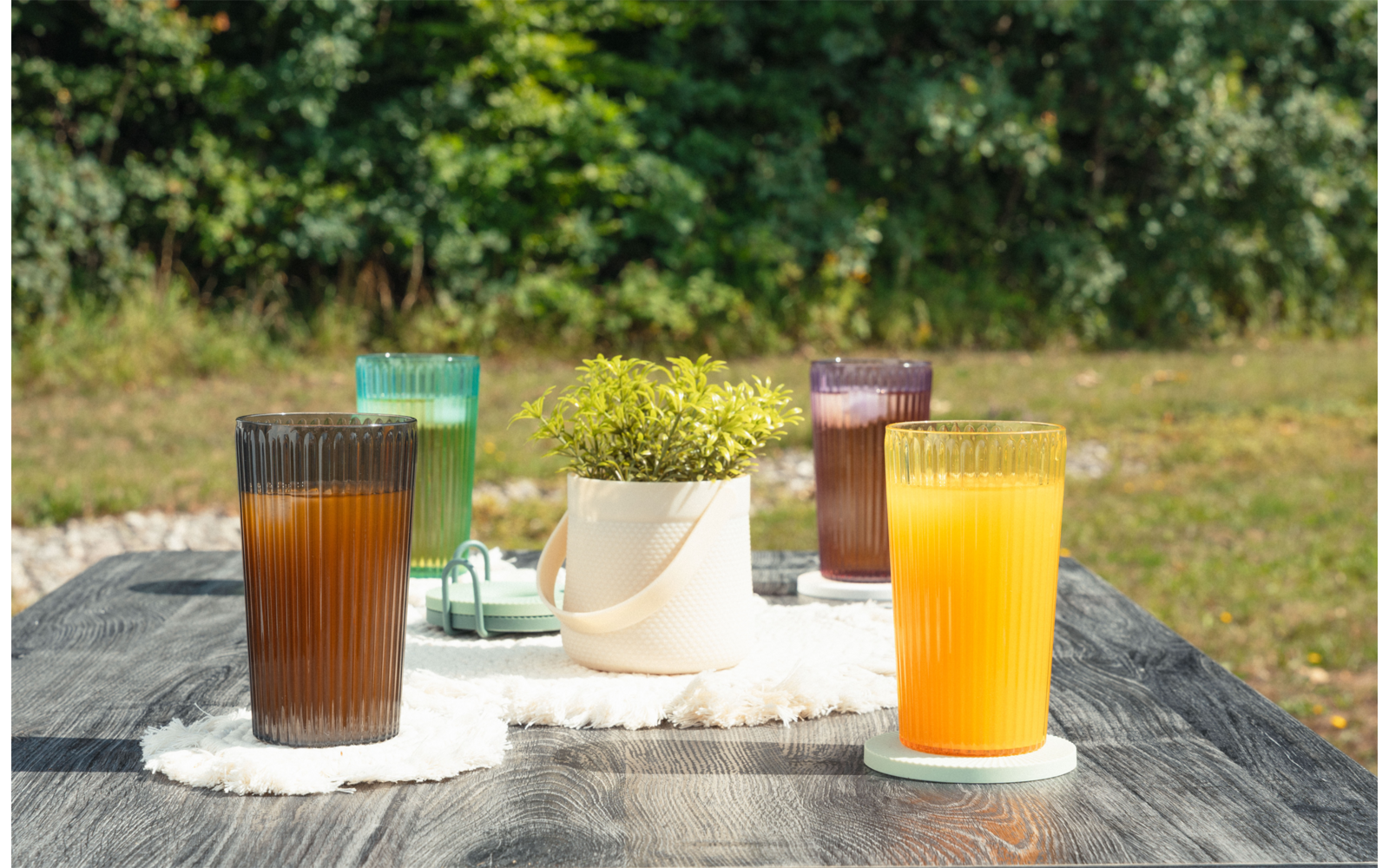 Mesa de madera gris al aire libre con cuatro vasos acanalados (marrón, verde, morado, naranja), una maceta blanca con planta verde sobre un paño blanco, césped y árboles al fondo