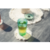 Vaso verde acanalado con líquido amarillo-verde y cubitos de hielo sobre un posavasos blanco de ganchillo sobre una mesa de madera oscura; al fondo un pequeño plato verde y una taza clara