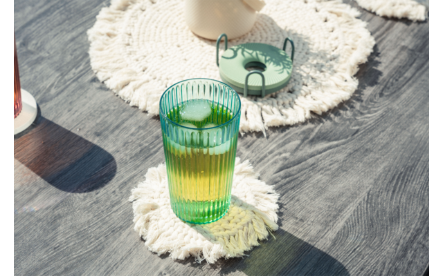 Vaso verde acanalado con líquido amarillo-verde y cubitos de hielo sobre un posavasos blanco de ganchillo sobre una mesa de madera oscura; al fondo un pequeño plato verde y una taza clara