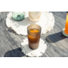Vaso marrón acanalado con líquido naranja sobre un posavasos redondo de macramé blanco en un suelo de madera gris; en el fondo se ven otros dos vasos sobre posavasos similares