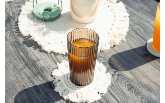Vaso marrón acanalado con líquido naranja sobre un posavasos redondo de macramé blanco en un suelo de madera gris; en el fondo se ven otros dos vasos sobre posavasos similares