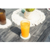 Vaso con líquido de color naranja sobre un posavasos en una mesa de madera oscura, un segundo vaso al fondo, un tapete blanco tejido y una maceta con planta verde