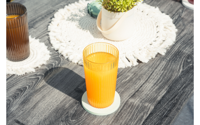 Vaso con líquido de color naranja sobre un posavasos en una mesa de madera oscura, un segundo vaso al fondo, un tapete blanco tejido y una maceta con planta verde