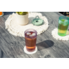 Dos vasos estriados con cubitos de hielo sobre posavasos blancos en una mesa de madera gris; al fondo una maceta blanca con una planta verde sobre un salvamanteles redondo de macramé y un pequeño recipiente metálico verde