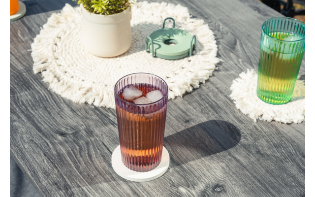 Dos vasos estriados con cubitos de hielo sobre posavasos blancos en una mesa de madera gris; al fondo una maceta blanca con una planta verde sobre un salvamanteles redondo de macramé y un pequeño recipiente metálico verde