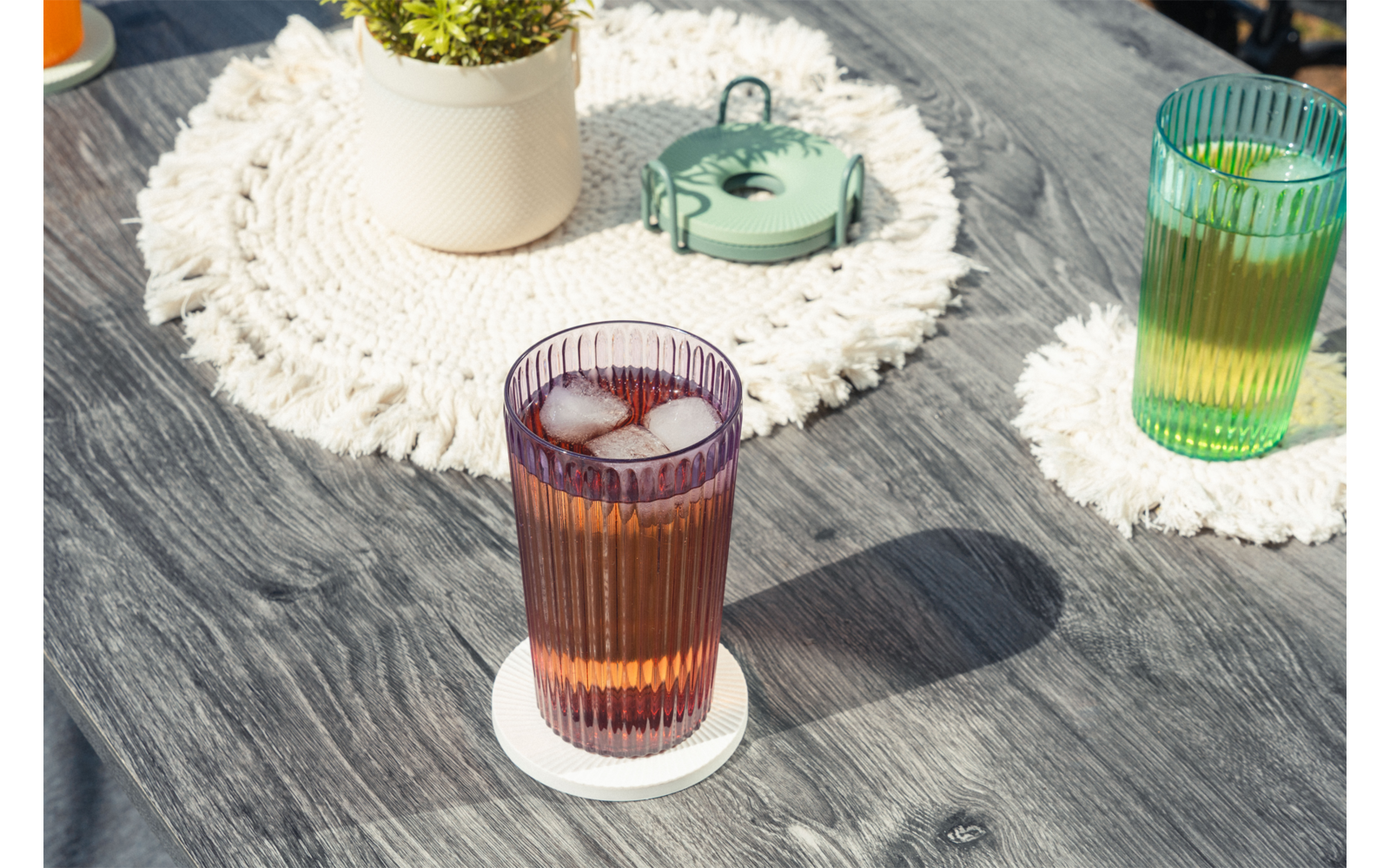 Dos vasos estriados con cubitos de hielo sobre posavasos blancos en una mesa de madera gris; al fondo una maceta blanca con una planta verde sobre un salvamanteles redondo de macramé y un pequeño recipiente metálico verde