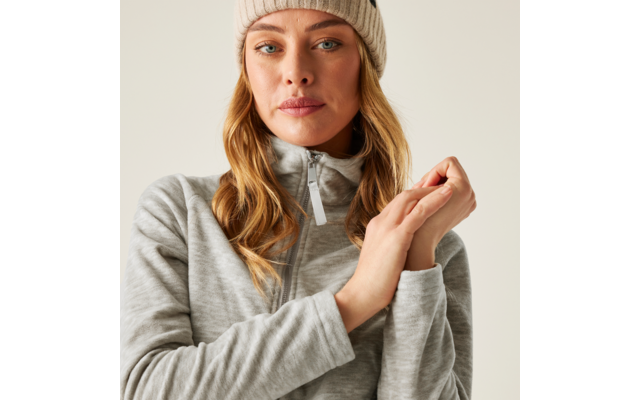 Nahaufnahme einer Frau in hellgrauem Zip-Sweater und beiger Strickmütze, Hände vor der Brust, neutraler Hintergrund
