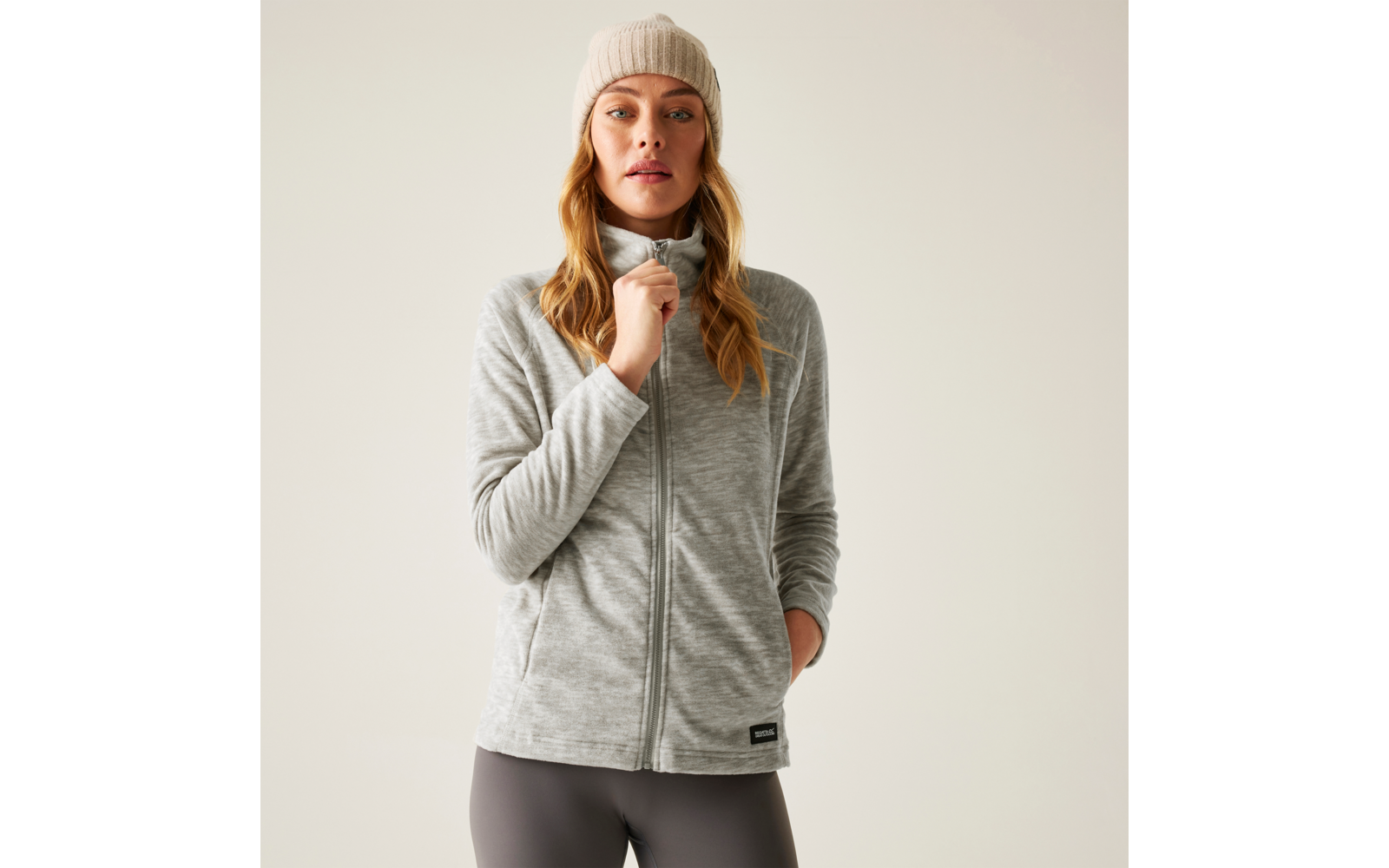 Person frontal stehend, trägt graue Fleecejacke mit Reißverschluss, beige Strickmütze, dunkle Leggings; eine Hand hält den Reißverschluss, andere Hand in Tasche, heller einfarbiger Hintergrund
