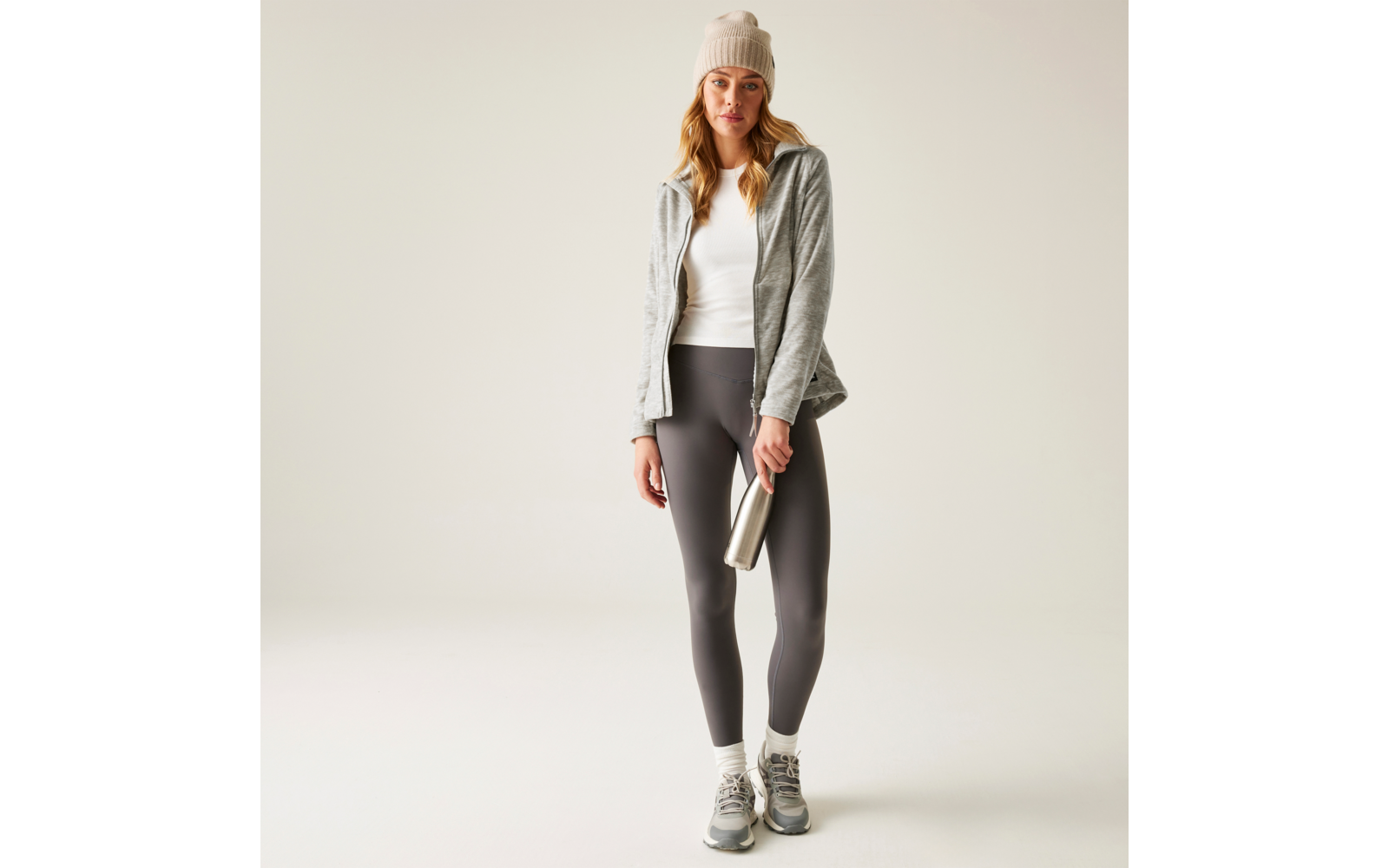 Person steht vor hellem Hintergrund, trägt graue Mütze, graue Jacke, weißes Top, dunkle Leggings, weiße Socken und graue Sneaker, hält eine silberne Trinkflasche in einer Hand