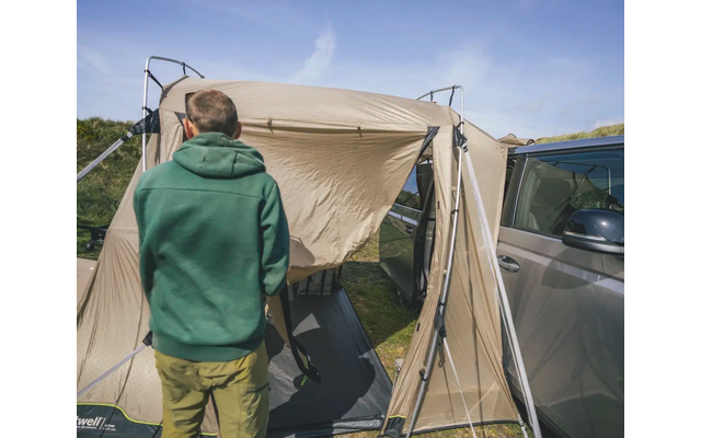 Person in grüner Jacke steht vor einem beigefarbenen Zelt neben einem grauen Auto unter blauem Himmel