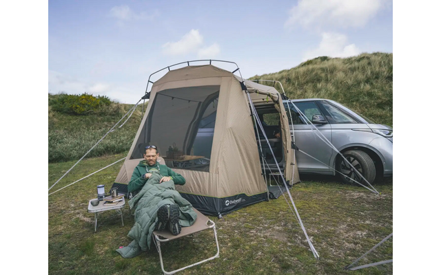 Person in grünem Anzug sitzt vor einem großen Zelt neben einem Auto in einer grünen Landschaft