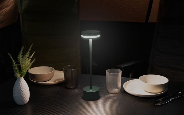 Eingeschaltete grüne Tischlampe in der Mitte eines dunklen Tisches; links eine weiße Vase mit grünem Zweig; links und rechts jeweils ein Teller mit einer Schale; zwei durchsichtige Gläser vor der Lampe; Messer und Gabel rechts