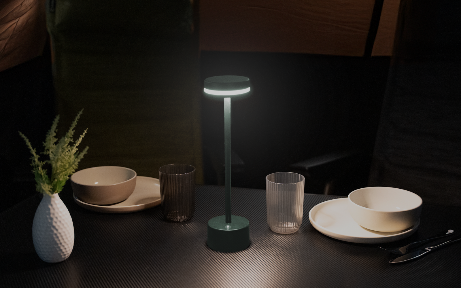 Eingeschaltete grüne Tischlampe in der Mitte eines dunklen Tisches; links eine weiße Vase mit grünem Zweig; links und rechts jeweils ein Teller mit einer Schale; zwei durchsichtige Gläser vor der Lampe; Messer und Gabel rechts